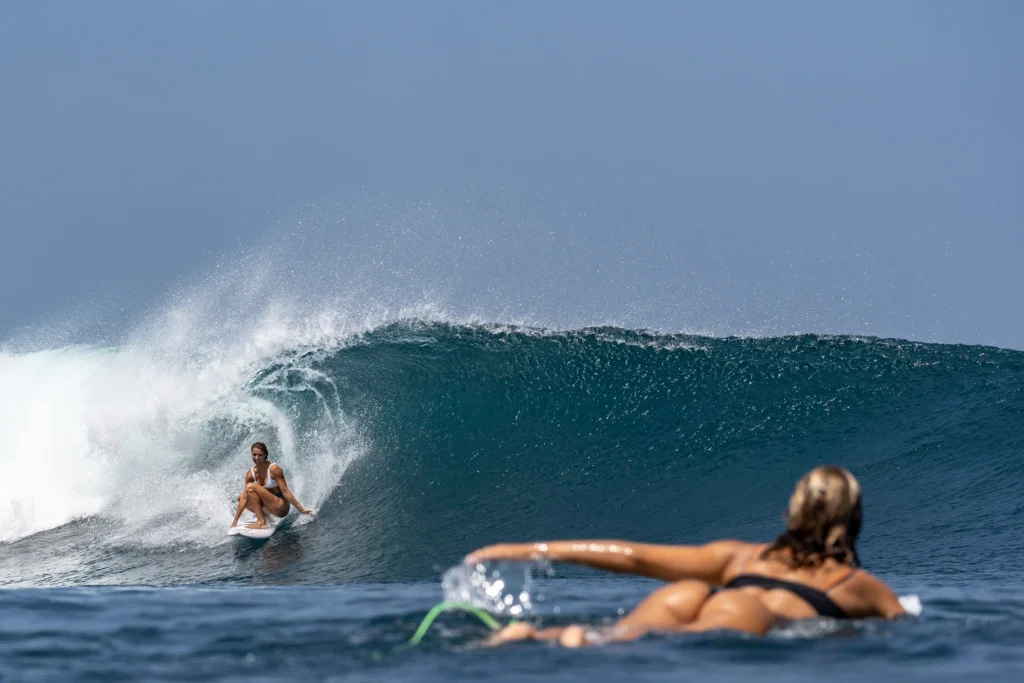 Female surfer riding a powerful blue wave while another surfer paddles in the foreground