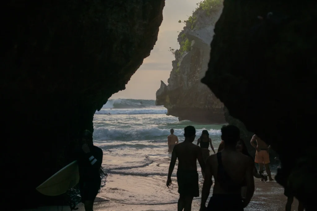 Group of surfers carrying surfboards through a rocky beach entrance toward the ocean in Bali